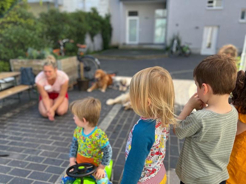 Drei Kinder schauen in eine Richtung, im Hintergrund sieht man eine Frau und einen Hund